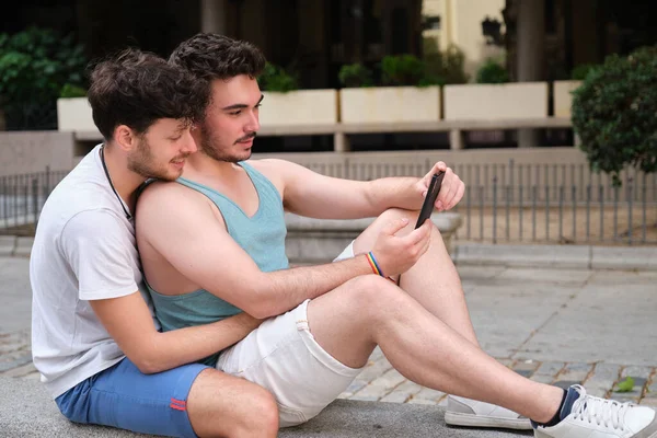 Γκέι ζευγάρι που χρησιμοποιεί το smartphone κάθεται σε ένα παγκάκι. — Φωτογραφία Αρχείου
