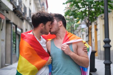 Eşcinsel bir çift bankta oturmuş gülüyor ve sarılıyor ellerinde LGBT bayrakları.