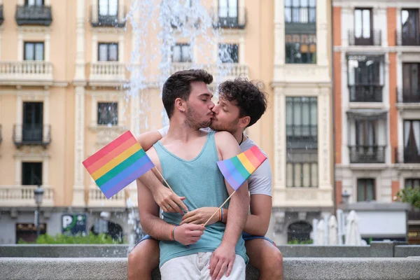 Cuplu gay râzând și îmbrățișându-se așezat pe o bancă care deține steaguri LGBT. — Fotografie, imagine de stoc