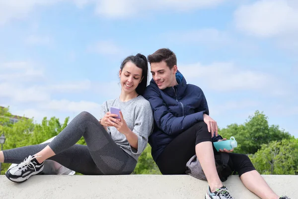 Feliz joven pareja deportiva comprobando en el teléfono inteligente mientras está sentado en un parque. — Foto de Stock