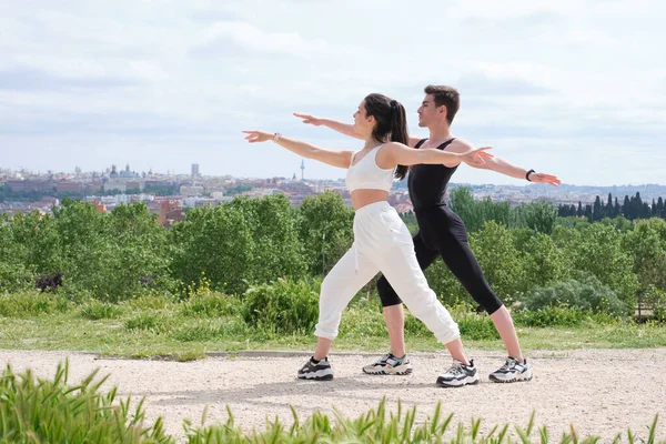 Young couple doing warrior two yoga pose in a park.