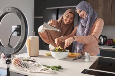 Modern mutfakta salataya zeytin yağı dökerken gülümseyen iki müslüman yemek vlogcusu..