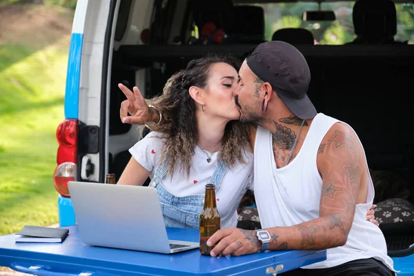 Young tattooed couple kissing in front of the laptop, sitting in a picnic table during a van trip.