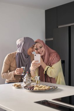 Mutfak masasında çay ve Arap tatlıları içerken gülüp akıllı telefon kullanan iki müslüman kadın..