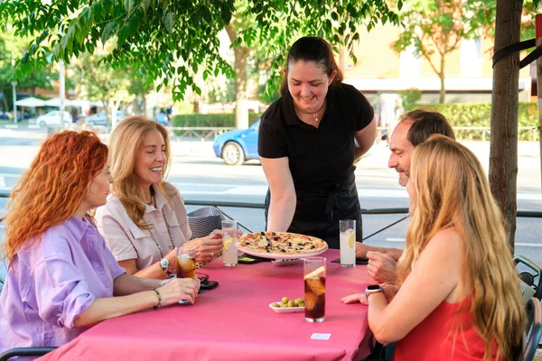 Waitress serving pizza to a group of mature friends at a restaurant table.