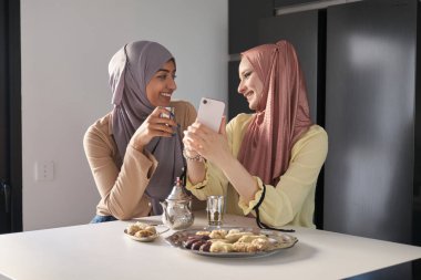 Mutfak masasında çay ve Arap tatlısı içerken akıllı telefon kullanan iki Müslüman kadın..