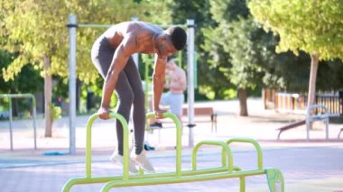 Young fit shirtless black man doing calisthenics workout on parallel bars outdoors on sunny day. Fitness and sport lifestyle.