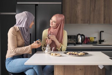 İki genç Müslüman kadın konuşuyor, çay içiyor ve mutfak masasında Arap tatlısı yiyorlar..