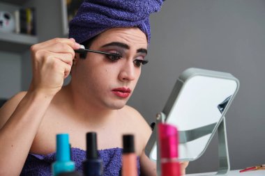 Young drag queen applying eye mascara after shower with a towel on his head.