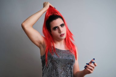 Portrait of drag queentaking off red wig and looking at camera on grey background. LGBTQ queer.