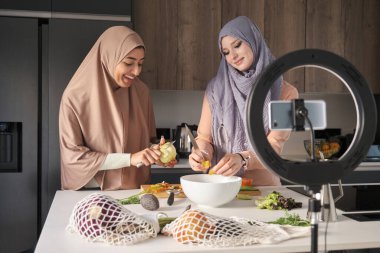 Two muslim content creators slicing, chopping and peeling vegetables at kitchen to prepare a salad.