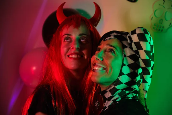 Two women dressed as a harlequin and devil smiling at a costume Halloween party.