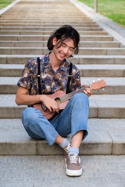 Genç Tayvanlı adam gülümseyip akustik gitar çalıyor. Merdivenlerde oturuyor..
