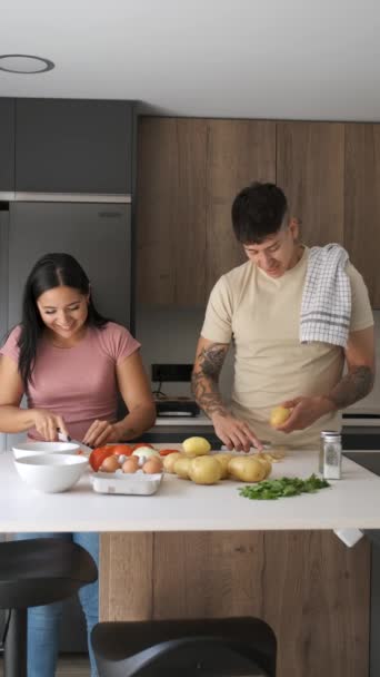 2人の若いラテン人が笑って ジャガイモを剥離しながら話して キッチンでレシピを準備するためにトマトを切る — ストック動画