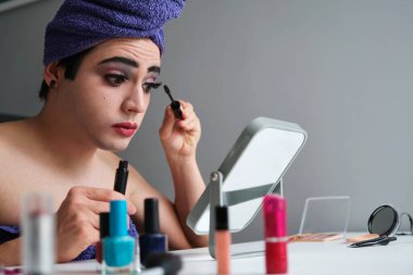 Genç transseksüel adam duş aldıktan sonra kafasında havluyla göz maskarası sürüyor..