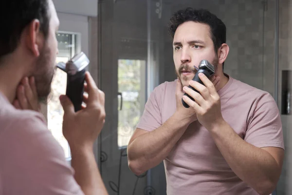 Young bearded man trimming his beard with a trimmer in bathroom.