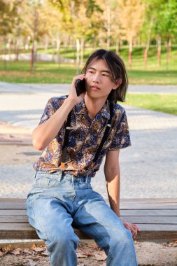 Genç Asyalı adam akıllı telefonda konuşuyor. Sokakta bankta oturuyor..