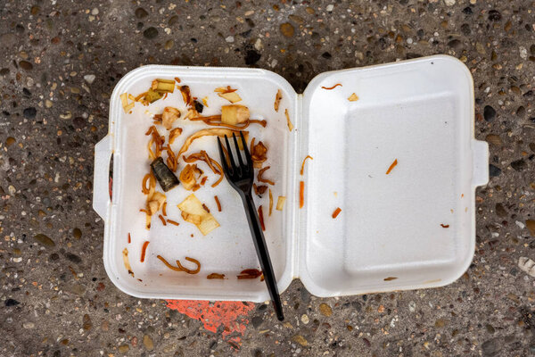 Discarded polystyrene container with remnants of a meal of takeaway food lying on the ground with a plastic disposable fork in a pollution and garbage concept