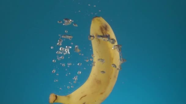 スローモーション新鮮なジューシーな1つの黄色のバナナは スプラッシュや泡で水に落ちます 青い背景の果物 — ストック動画