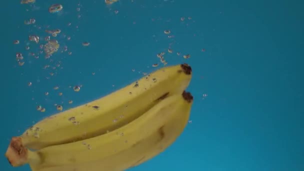 スローモーション新鮮なジューシーな2つの黄色のバナナは スプラッシュや泡で水に落ちます 青い背景の果物 — ストック動画