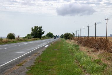 Yeşil çimenlerin, dikenli kahverengi bitkilerin, kablolu elektrik desteklerinin ve mavi bulutlu gökyüzünün olduğu yol kenarına bakın. Turistler için yol turu iyi bir deneyimdir..