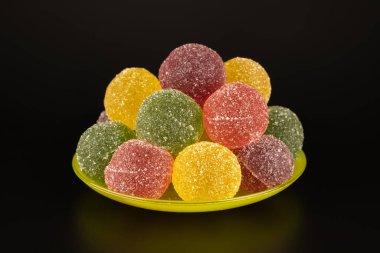 Siyah arka plandaki çay tabağındaki renkli tatlılar marmeladı. Meyve jölesi, sarı, kırmızı, pembe, üzerine şeker serpiştirilmiş. Yemek konsepti, lezzetli tatlı yiyecekler..