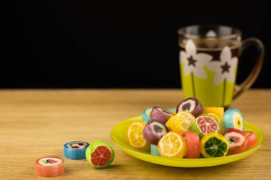 Ahşap desenli arka planda meyveler çizilmiş renkli karamel şekerler. Bir fincan aromatik kahve ve tatlı. Yemek konsepti, lezzetli tatlı yiyecekler..