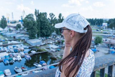 Woman in white cap thoughtfully looks at picturesque river and motor boats parked. She stands on the bridge leaning on the iron-concrete handrail. Garage system for speedboats.