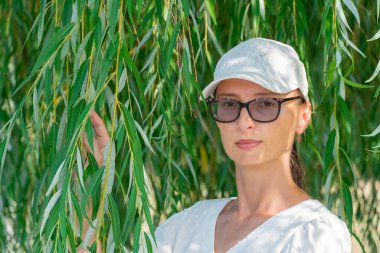 Close-up woman the background of willow. Hand holds green branches tree. She is wearing white blouse and cap. Romantic portrait of woman. Summertime.
