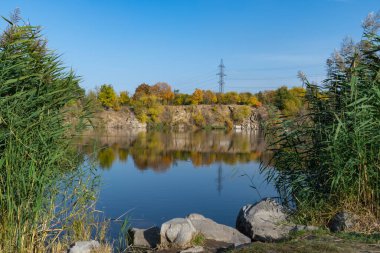 Autumn colors in sunny day on lake in city park. Landscape golden foliage and reflection of trees in calm water. Colorful plants on stone bank of pond and blue sky. Autumn reflections. Fall bright.