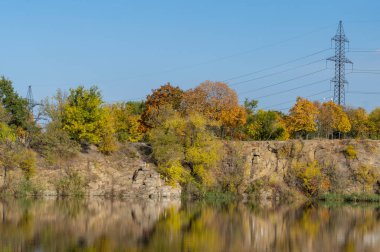 Autumn colors in sunny day on lake in city park. Landscape golden foliage and reflection of trees in calm water. Colorful plants on stone bank of pond and blue sky. Autumn reflections. Fall bright.