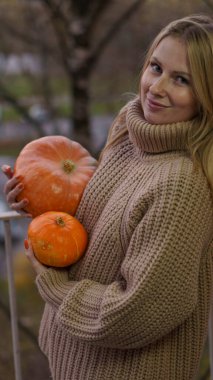 Yakından bakınca gülümseyen sarışın kız elinde iki tane olgun turuncu balkabağı tutuyor. Kız bej örülü bir kazak giyiyor.