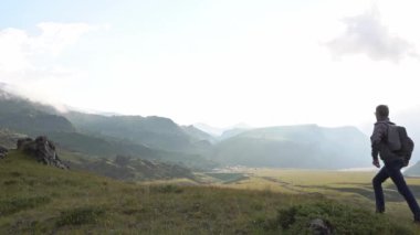 a young man tourist walks along the gentle slope of the mountain and, stopping, raises his hands, rejoicing and enjoying the landscape.  summer day, close-up.  Caucasus Mountains, Elbrus 