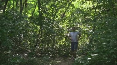 a young man in a white meek and gray shorts with headphones runs through the forest on a summer morning. the bright sun shines, hard shadows.