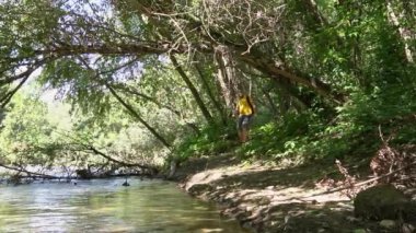 a young man traveler in a yellow T-shirt and a large backpack descends from the forest to the river. the bright sun is shining. hard shadows. overall plan