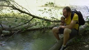 a young man traveler with a beard in a yellow T-shirt and a backpack sits on a fallen tree near the river and eats.  medium plan.  bright sun, harsh shadows