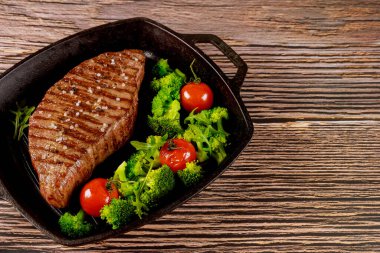 Orta pişmiş biftek, brokoli ve ahşap arka planda ızgara tavada domates..