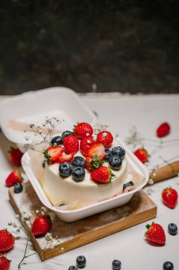 Çilek, yabanmersini ve çiçeklerle süslenmiş güzel beyaz pasta, beyaz arka planda tahta bir tahtanın üzerinde duruyor.