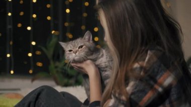 Ekose gömlekli genç bir kadın İskoç kedisini elinde tutuyor ve öpüyor.