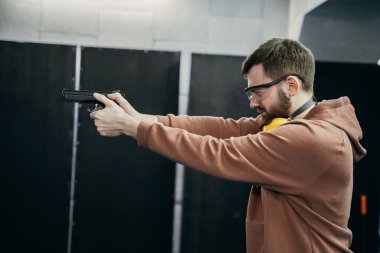 Bir adam ateş etmek için doğru pozisyonda duruyor ve hedefi vurmaya hazırlanıyor, elinde bir tabanca tutuyor, atış poligonuna ateş ediyor.