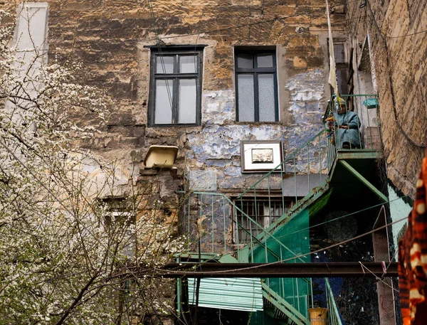 Odessa, Ukraine - 04 18 21: typical old Odessa Kanava street courtyard. And old lady sitting on the stairs in front of scratched textured building facade. Spring blossom time — Fotografia de Stock