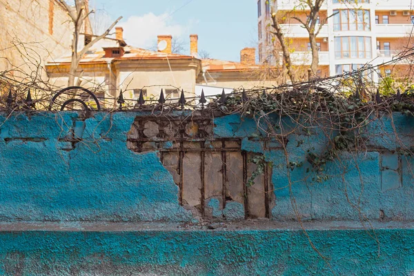 Rusty forged metal hedge visible through a crack in plastered blue painted fence. Old house roof and new residential building building on the background. A photo with copy space for text — Fotografia de Stock