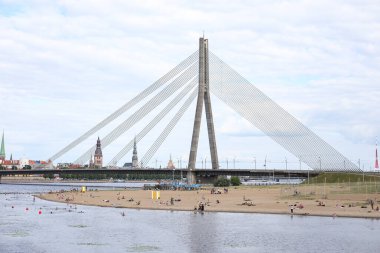 Riga şehrinin manzarası eski asma köprü ve arka planda eski mimari ve Daugava nehri yakınlarındaki kum plajı. Fotoğraf sıcak ve bulutlu bir yaz gününde çekildi..