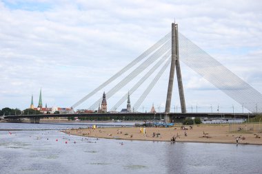 Riga şehrinin manzarası eski asma köprü ve arka planda eski mimari ve Daugava nehri yakınlarındaki kum plajı. Fotoğraf sıcak ve bulutlu bir yaz gününde çekildi..