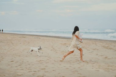 Kadın ve köpek koşuyor ve sahilde yakalamaca oynuyorlar.