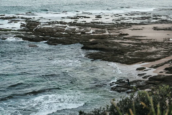 Gentle waves breaking on rocks at the beach with man fishing in the distance