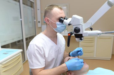 Dişçi koltuğunda oturan genç bir kız ve yanında oturan bir dişçi. Diş mikroskobuyla dişlere bakıyor ve elinde bir diş fırçası ve ayna tutuyor. İşlem sırasında dişçi ve hasta