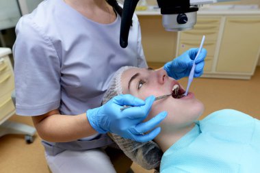 Genç bir dişçi uzmanı mikroskop kullanarak modern beyaz diş hekimliğindeki güzel bir kadının dişlerini muayene eder ve tedavi eder. Diş protezi, tedavi ve diş beyazlatma..