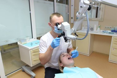 Erkek dişçi genç ve güzel bir kadını muayene eder dişlerine cerrahi diş muayenehanesinde profesyonel bir mikroskopla bakar.