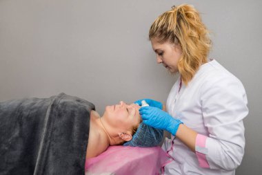 Profesyonel bir kozmetikçi, tüpten kanepeye uzanmış bir kadının yüzüne krem sürer..